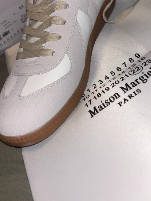 Maison Margiela vita sneakers - Maison Margiela sneakers i vitt och beige med klassisk gummisula. Skon har en stilren design med mockadetaljer och vita paneler, samt beige snören. Perfekt för dig som gillar minimalistisk och trendig stil.