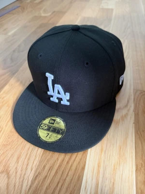 Svart LA New Era keps - Snygg svart keps från New Era med LA-logga broderad framtill i vitt. Klassisk 59FIFTY-modell med platt skärm och broderade ventilationshål. MLB-logga baktill. Perfekt för dig som gillar streetwear och sportig stil.