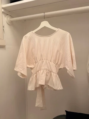Vit omlottopp med knytband - Säljer en luftig vit omlottopp med knytband i midjan och korta, vida ärmar. Toppen har en avslappnad passform och volangdetalj nedtill. Perfekt för varma dagar och enkel att styla med olika outfits.