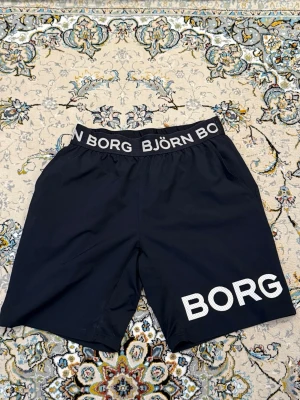 Svarta Björn Borg tränings shorts med logga - Snygga svarta shorts från Björn Borg med bred resår i midjan där märket står tryckt runtom. Stora vita BORG-bokstäver på ena benet. Perfekta för träning eller chill. Klassisk passform och sportig look.