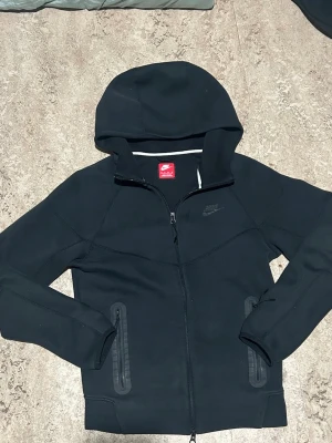 Svart Nike Tech Fleece set - Nike Tech Fleece set i svart med hoodie och matchande byxor. Jackan har dragkedja, huva och två fickor med dragkedja. Byxorna har dragsko i midjan och dragkedjefickor. Perfekt för träning eller chill. Snygg och stilren design med diskret Nike-logga. Tröjan st Xs och byxorna st S