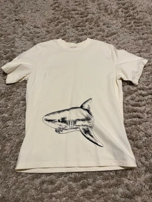 Vit Palm Angels t-shirt med hajmotiv - Säljer en vit t-shirt från Palm Angels med ett stort svart hajmotiv både fram och bak. På ryggen står det 'Palm Angels' i gotisk stil. T-shirten har klassisk passform och rund hals. Perfekt för dig som gillar streetwear och unika prints.