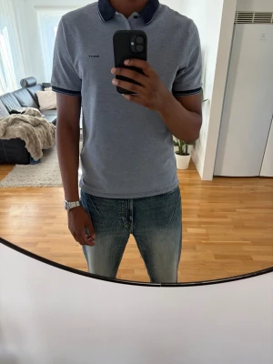 Blå pikétröja med mörkblå krage - Säljer en stilren blå pikétröja med mörkblå krage och ärmslut. Tröjan har knappar vid halsen och diskret logga på bröstet. Perfekt till jeans för en avslappnad look. Även fin blå kontrast mellan tröja och krage. 