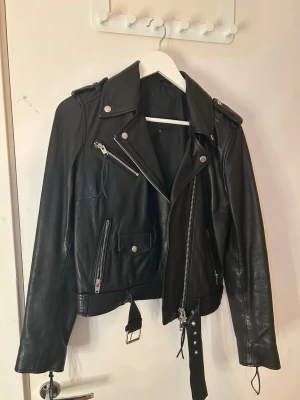 Svart skinnjacka Rockandblue 38 - Snygg svart skinnjacka från Rockandblue i storlek 38. Klassisk bikerstil med silvriga dragkedjor, axelklaffar och bälte i midjan. Jackan har flera fickor med dragkedja och tryckknappar för extra detaljerad look. Använd fåtal gånger ( nyskick )