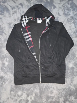 Svart hoodie med dragkedja från Burberry - Snygg svart hoodie från Burberry med dragkedja och klassiskt rutigt foder i huvan. Justerbar huva med snören och två fickor framtill. Broderad logga på bröstet och mjukt material som känns skönt mot huden. Perfekt för en avslappnad och trendig look. Den passar lite stor.