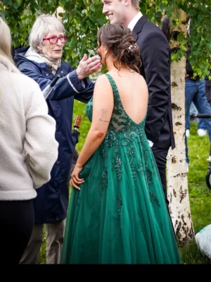 Grön långklänning med spets och v-ringning -  inga tecken på användning i varken kjolen eller toppen. Inga pärlor har fallit av men skickar med exakta pärlor som kom med klänningen. Kan även skicka med flugan och näsduken som är samma färg som klänningen om det skulle vara intressant. mått i näst sista bilden. dock sytt in klänningen på sidan under armarna lite (vilket jag visar på sista bilden), går att sprätta upp om köparen vill de. Går att sys in endel om måtten inte stämmer så som jag gjorde. som den är nu är den i str M ungefär. 