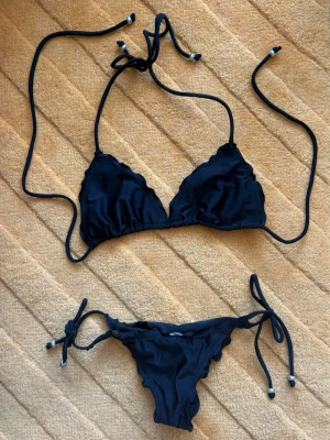 Wonderland svart bikini med knyt, volang & scrunch - Snygg svart bikini med triangelformad topp och knyt både i nacken och ryggen. Underdelen har knyt i sidorna, scrunch bak för en smickrande figur och fina volangdetaljer längs kanterna. Perfekt för lata dagar på stranden eller poolen. Oanvänd. Strl M (topp) & XS (trosa). Se gärna mina övriga annonser för bundlerabatter och samfrakt!