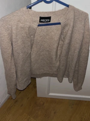 Beige kofta från PIECES - Mysig beige kofta från PIECES med rund halsringning och knappar framtill. Koftan har en enkel, rak passform och är stickad i ett mjukt material. Perfekt att slänga över en t-shirt eller topp för en avslappnad look.
