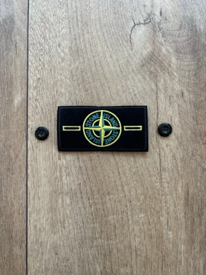 Stone Island Original Patch / Badge – Helt ny & oanvänd - Säljer en klassisk Stone Island-badge med tillhörande originalknappar, precis som visas i bilden. Patchen är helt ny, aldrig använd och i perfekt skick. Perfekt som reservdel till din Stone Island-jacka eller tröja. Klassisk gul/grön design. 
