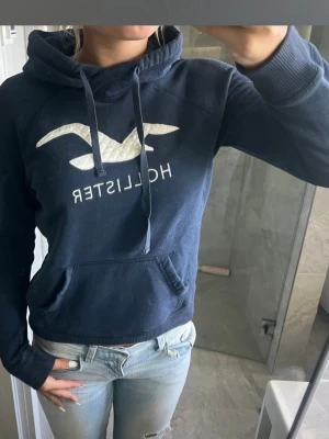 Mörkblå hoodie från Hollister - Mörkblå hoodie från Hollister med stor vit logga och fågeltryck på bröstet. Klassisk känguruficka framtill, dragsko i huvan och ribbade muddar. Perfekt för en avslappnad och cool stil.