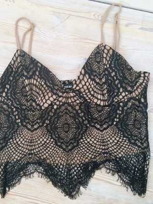 Topp med svart spets - Snyggt linne/topp med beige bas och svart spetsmönster. Toppen har smala axelband och v-ringad hals, samt dekorativ spetskant nedtill. Perfekt för dig som vill ha en trendig och feminin look.