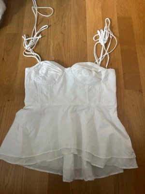 Vit korsettopp med volang från ZARA - Supersnygg vit korsettopp från ZARA med tunna axelband och volang nedtill. Toppen har markerade kupor och snörning i ryggen för en extra snygg passform. Perfekt för dig som vill ha en trendig och feminin look.