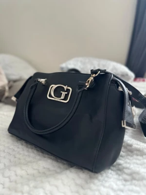 Svart handväska från Guess - Snygg svart handväska från Guess med silverfärgad logga framtill. Väskan har dubbla handtag och avtagbar axelrem. Tillverkad i ett slätt konstläder med stilren design och metalliska detaljer. Perfekt för dig som vill ha en trendig och praktisk accessoar.