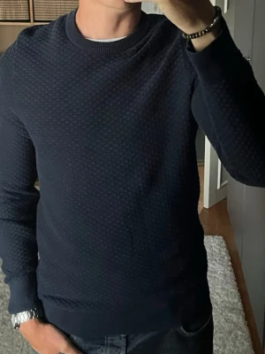J.Lindeberg stickad tröja i mörkblå - stl M - Tätt stickad med små rutor i mönstret och ribbad hals, muddar och nederkant. Rak passform, skön under jacka när det blir svalare. Personen på bilden är 189cm, väger 78kg.  Använd men i mycket fint skick, inga hål eller fläckar.  Rök- och djurfritt hem. Skriv gärna om du vill ha fler bilder eller undrar något!