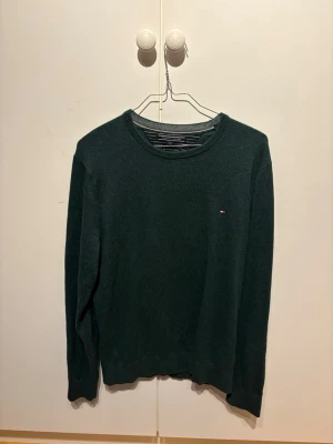 Mörkgrön stickad tröja Tommy Hilfiger - Mörkgrön stickad tröja från Tommy Hilfiger i bomull och kashmir. Klassisk rund halsringning och liten broderad logga på bröstet. Perfekt till lager på lager och enkel att matcha med jeans eller chinos.