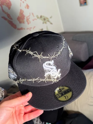 Svart keps med taggtråd och skelett brodyr  - Säljer en svart 59FIFTY keps med broderat taggtrådsmönster och vit 'Sox'-logga framtill. På sidan finns detaljerade broderier av skelett med vapen och rosor. Platt skärm och klassisk snapback-stil. Perfekt för dig som gillar streetwear och unika detaljer.