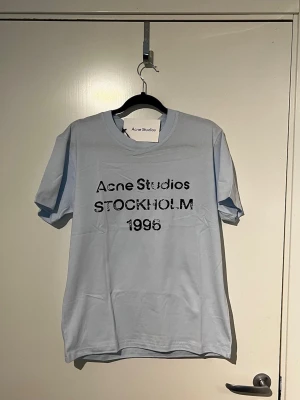 Ljusblå Acne Studios t-shirt - Säljer en ljusblå t-shirt från Acne Studios med trycket 'Acne Studios STOCKHOLM 1996' i svart på bröstet. Klassisk rund hals och korta ärmar. Perfekt för dig som gillar stilrena plagg med en twist.