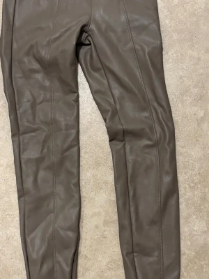 Ljus brun  leggings i fuskläder - Snygga bruna leggings i fuskläder med raka ben och dekorativa sömmar längs framsidan. De har en dragkedja vid ankeln och resår i midjan för extra komfort. Perfekta för dig som vill ha en trendig och edgy look.