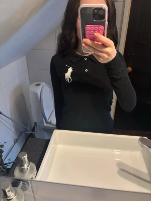 Svart långärmad piké från Polo Ralph Lauren - Säljer en svart långärmad pikétröja från Polo Ralph Lauren med klassisk krage och knappar framtill. Stor vit broderad logga på bröstet ger en clean och sportig look. Perfekt till jeans eller kjol för en stilren outfit.