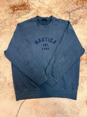 Blå sweatshirt från Nautica - Säljer en blå sweatshirt från Nautica med trycket 'NAUTICA EST. 1983' på bröstet. Tröjan har rund halsringning, långa ärmar och ribbade muddar vid ärmslut och nederkant. Perfekt för en avslappnad och stilren look.