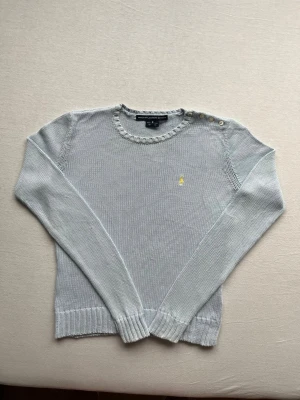 Stickad Ralph Lauren tröja - Ljusgrå stickad tröja med rund halsringning och dekorativa knappar på axeln. Tröjan har ett gult broderat märke på bröstet och ribbade muddar vid ärmslut och nederkant. Långärmad och i mjukt material, perfekt för lager på lager.