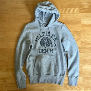 Grå Hilfiger Denim hoodie med tryck - Snygg grå hoodie från Hilfiger Denim med stort tryck på bröstet och klassisk känguruficka. Tröjan har huva med dragsko och ribbade muddar vid ärmar och nederkant. Perfekt för en avslappnad och cool stil.