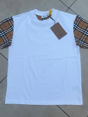 Vit t-shirt med rutiga ärmar - Snygg vit t-shirt med klassiska beige, svarta och röda rutiga ärmar. T-shirten har rund halsringning och normal passform. Perfekt för dig som vill ha en stilren look med en twist. Tillverkad i mjuk bomull för skön känsla.