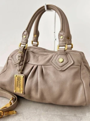 Beige handväska i skinn med gulddetaljer - Snygg beige handväska i mjukt skinn med dubbla handtag och avtagbar axelrem. Väskan har guldfärgade metalldetaljer och dekorativ bricka framtill. Klassisk form med veckad framsida och detaljerade sömmar.