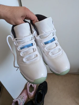 Vita Air Jordan 11 sneakers - Säljer ett par vita Air Jordan 11 sneakers med blå detaljer och genomskinlig grönaktig sula. Skorna har högt skaft, vita skosnören och en ikonisk Jumpman-logga på sidan. Ovandelen är i textil och lackat läder för en clean look. Använda 2 gånger.
