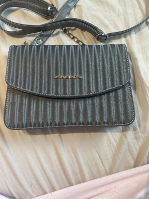 Grå axelväska från Michael Kors - Snygg grå axelväska från Michael Kors i konstläder med räfflad struktur och guldfärgad logga framtill. Väskan har justerbar axelrem och ett rymligt huvudfack med innerficka för småsaker. Perfekt för dig som vill ha en stilren och trendig väska.