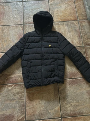 Svart Lyle&Scott jacka med huva - Säljer en svart Lyle&Scott jacka med huva och dragkedja framtill. Jackan har två sidofickor och ett gult broderat logomärke på bröstet. Tillverkad i vadderat material som håller värmen under kalla dagar. Perfekt för vintern och hösten.