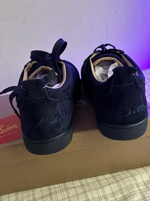 Christian Louboutin, ny  - Stilrena svarta sneakers från Christian Louboutin med klassisk röd sula och svarta skosnören. Skorna har en låg profil och är tillverkade i mocka med detaljer i läder. Perfekta för dig som vill ha en exklusiv och trendig look.