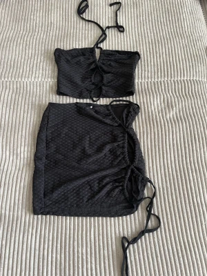 Svart tvådelat set med struktur - Säljer ett svart tvådelat set med crop top och kjol i ett strukturerat tyg. Toppen har knytband framtill och kjolen har snörning i sidan för justerbar passform. Perfekt för dig som gillar trendiga och bekväma underkläder.