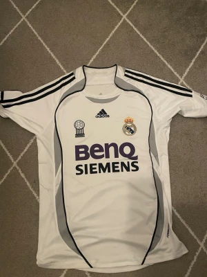 Real Madrid matchtröja Adidas S - Säljer en vit Real Madrid matchtröja från Adidas med svarta och grå detaljer. Tröjan har klubbmärke, Adidas-logga och BenQ Siemens sponsortryck på bröstet. Klassisk design med svarta ränder på axlarna och rund hals. Perfekt för fotbollsfans!