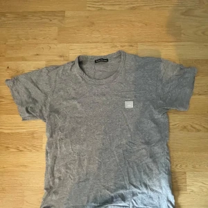 Grå t-shirt från Acne Studios - Snygg grå t-shirt från Acne Studios med klassisk rund hals och korta ärmar. T-shirten har den ikoniska Face-loggan broderad på bröstet och är gjord i mjuk bomull. Perfekt basic-plagg med clean look och avslappnad passform.