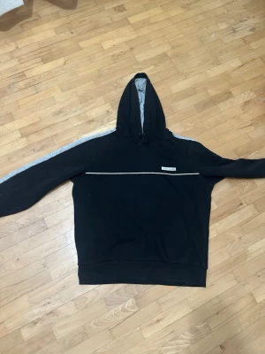 Svart hoodie från Jack & Jones - Svart hoodie från Jack & Jones med grå insida i huvan och gråa ränder längs ärmarna. Framtill finns en vit dekorativ linje och en liten logotyp på bröstet. Klassisk passform och mjukt material, perfekt för en avslappnad stil, passar både S och M  priset kan diskuteras 