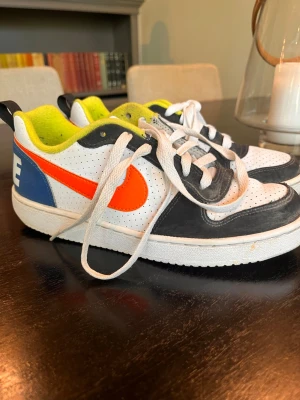 Nike vita sneakers med orange swoosh - Nike sneakers i vitt med svart och blåa detaljer, samt en stor orange swoosh på sidan. Insidan är neongul och ovandelen är perforerad för extra ventilation. Klassisk låg modell med vita snören och slitstark gummisula.