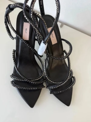 Svarta pumps med strassdetaljer Nelly - Snygga svarta pumps från Nelly med spetsig tå och tunna remmar dekorerade med glittrande strass. Klackarna har en elegant design och remmarna snirklar sig runt foten för en trendig look. Perfekta för att lyfta vilken outfit som helst. Klackarna är helt oanvända och köpa för 599kr. Jag är öppen för prisförslag🥰❤️