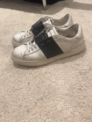 Valentino Garavani vita sneakers - Pris kan diskuteras!!!! Säljer ett par vita sneakers från Valentino Garavani med en bred svart läderdetalj över snörningen. Skorna har klassisk låg profil, vita snören och en vit sula. Tillverkade i läder och har en stilren, modern look.