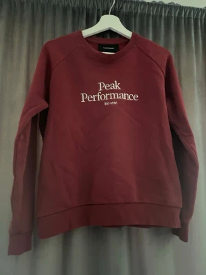 Vinröd sweatshirt Peak Performance - Snygg vinröd sweatshirt från Peak Performance med vit logga tryckt på bröstet. Klassisk rund halsringning och ribbade muddar vid ärmslut och nederkant. Perfekt för en avslappnad och sportig stil.