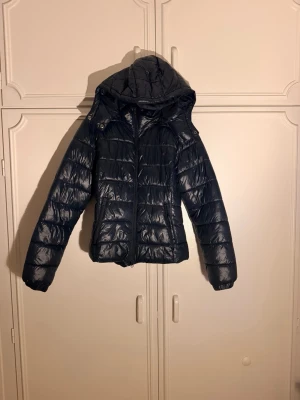 Mörkblå puffer vinterjacka  - Snygg svart dunjacka med glansig finish och avtagbar huva. Jackan har dragkedja framtill, två sidofickor och quiltad design som ger en modern look. Perfekt för kalla dagar när du vill hålla värmen och ändå se stilren ut.