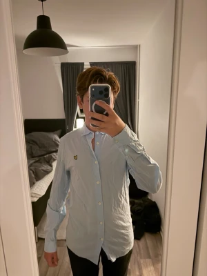 Ljusblå skjorta med knappdetaljer - Snygg ljusblå skjorta med klassisk krage och vita knappar. På bröstet finns en broderad gul logga som ger en extra touch. Skjortan har lång ärm och är tillverkad i bomull, perfekt för en clean och stilren look.