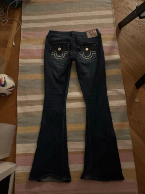  Mörkblå utsvängda, lågmidjade true religion jeans - Säljer ett par mörkblå lågmidjade, utsvängda true religion jeans med vit söm och guldknappar där bak, är i jättebra skick och jättebra passform, perfekt till dig som är ute efter snygga jeans!