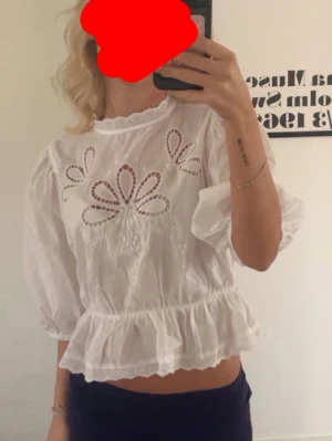 Vit blus med broderade detaljer - Söt vit blus med broderade blommönster och hålbroderier framtill. Blusen har volangkant nedtill, puffiga trekvartsärmar och rund hals med spetskant. Perfekt för en trendig och romantisk look.
