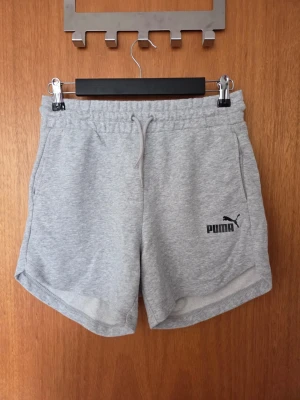 Grå sweatshorts från Puma - Snygga grå sweatshorts från Puma med elastisk midja och dragsko. Shortsen har två sidofickor och en liten Puma-logga i svart på vänster ben. Tillverkade i mjukt sweatshirtmaterial som är perfekt för chill eller träning.