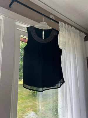 Svart ärmlös linnetopp med pärldetaljer - Snygg svart ärmlös linnetopp med rund hals och dekorativa pärldetaljer runt ringningen. Toppen har en lätt transparent känsla och en liten öppning bak i nacken för extra stil. Perfekt att matcha med jeans eller kjol.