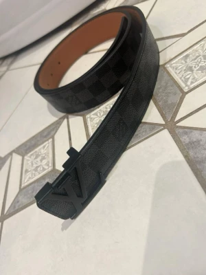 Svart rutigt bälte - Snyggt svart bälte med diskret rutigt mönster och en stilren logga som spänne. Bältet har en klassisk design och passar perfekt till jeans eller kostymbyxor. Insidan är ljusbrun vilket ger en snygg kontrast.