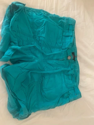 Turkosa shorts med fickor - Snygga turkosa shorts med flera stora fickor både fram och bak, shortsen har dragkedja och knapp framtill