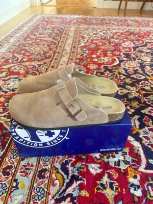 Birkenstock Boston mud  - Säljer ett par klassiska Birkenstock tofflor i beige mocka med justerbart spänne och korksula i storlek 43 . Innersulan är mjuk för extra skön känsla.