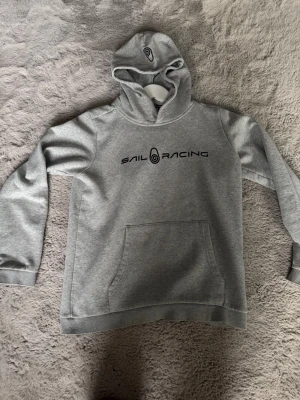 Grå hoodie från Sail Racing - Snygg grå hoodie från Sail Racing med klassisk huva och diskret logga på baksidan av huvan. Tillverkad i mjukt sweatshirtmaterial som känns skönt mot huden. Perfekt för en avslappnad och sportig stil.